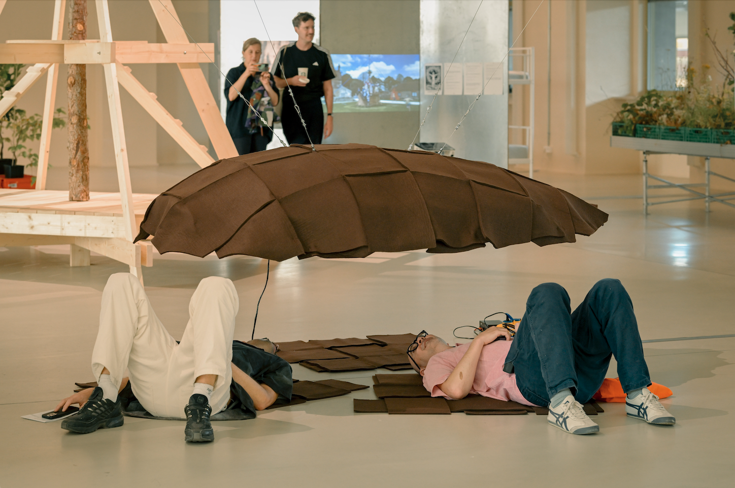 Zwei Besucherinnen einer Kunstaustellung liegen unter einem mit Filz und Holz gebauten, halbierten Apfel-Samen.